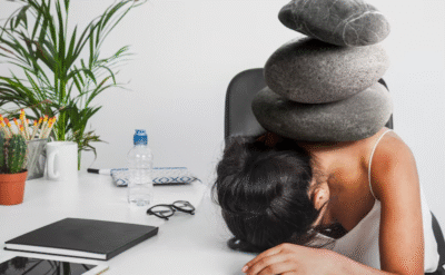 Stressed woman with stones on her back at work desk — symbolic of burnout in self transformation articles on The Power Within.