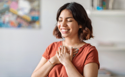 Smiling young woman with short hair holding her hands over her heart, eyes closed, expressing gratitude and inner peace in a cozy indoor setting.
